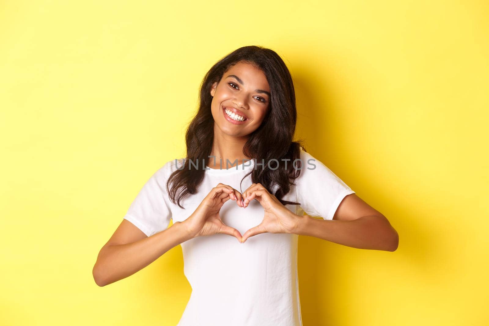 Image of lovely african-american girl in white t-shirt, showing heart sign and smiling, standing over yellow background.
