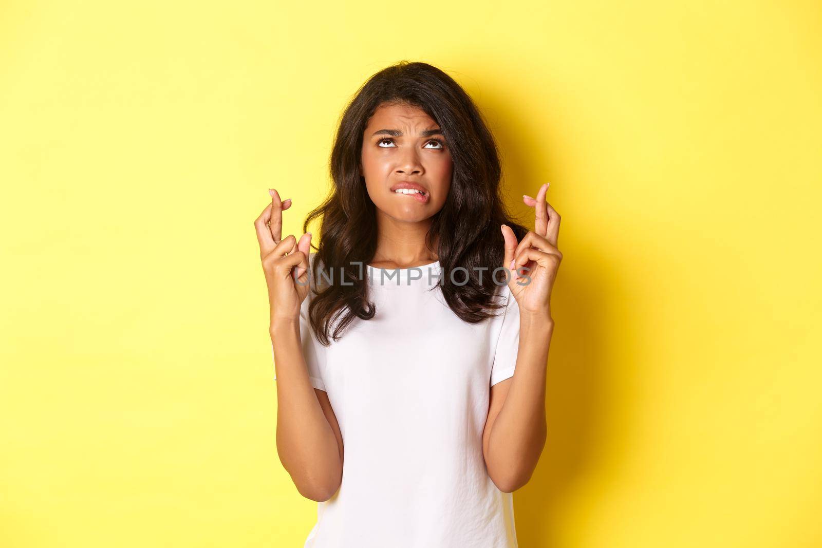 Portrait of worried and hopeful african-american girl, begging for something with fingers crossed, looking up and making wish, standing over yellow background.