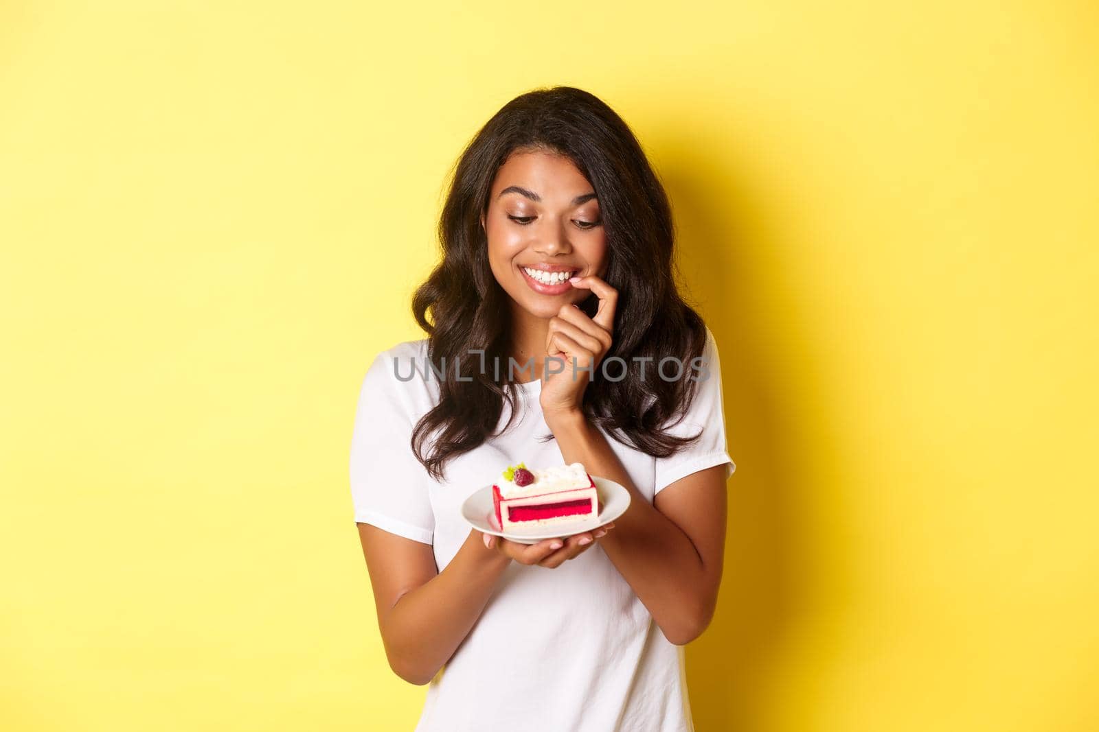 Image of beautiful african-american woman, looking with tempted smile at delicious piece of cake, standing over yellow background.