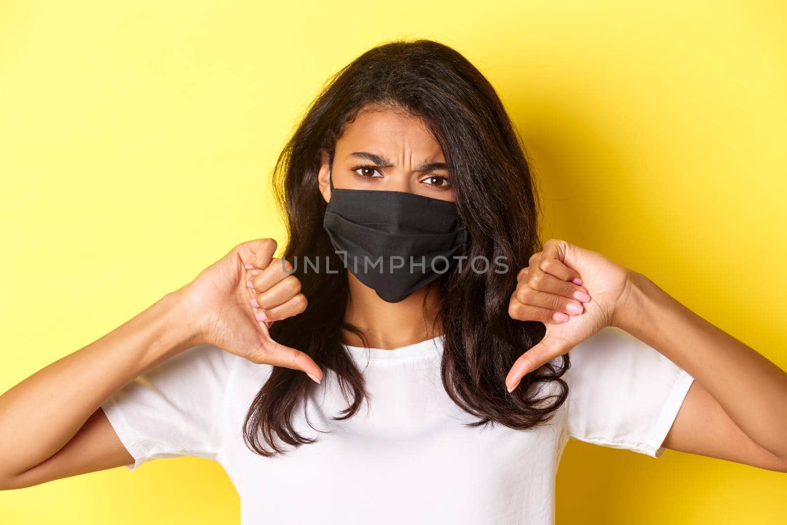 Concept of covid-19, social distancing and lifestyle. Close-up of displeased african-american girl in face mask, frowning upset and showing thumbs-down to disagree, yellow background by Benzoix