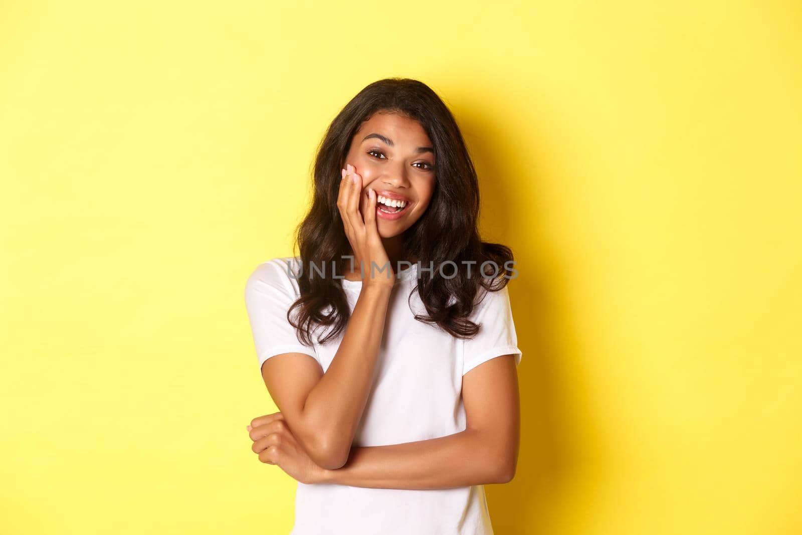 Portrait of attractive african-american girl, looking amazed and smiling, standing over yellow background in casual white t-shirt.