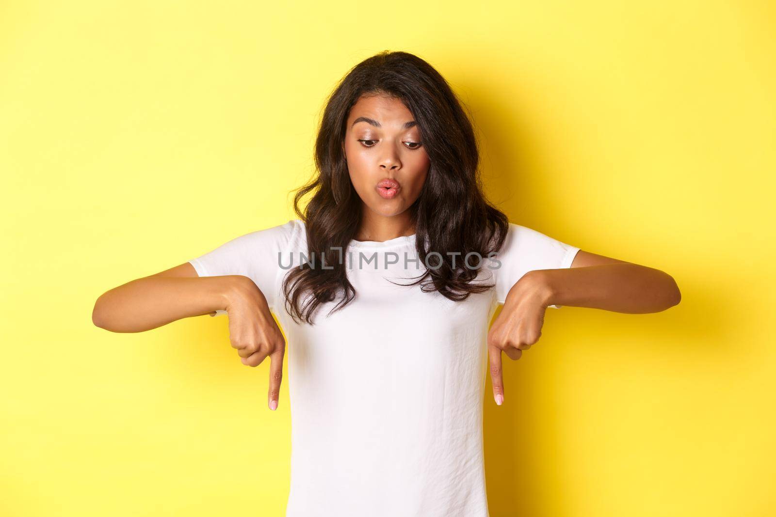 Portrait of amused, beautiful african-american female model in white t-shirt, saying wow while looking and pointing fingers down at something interesting, standing over yellow background.