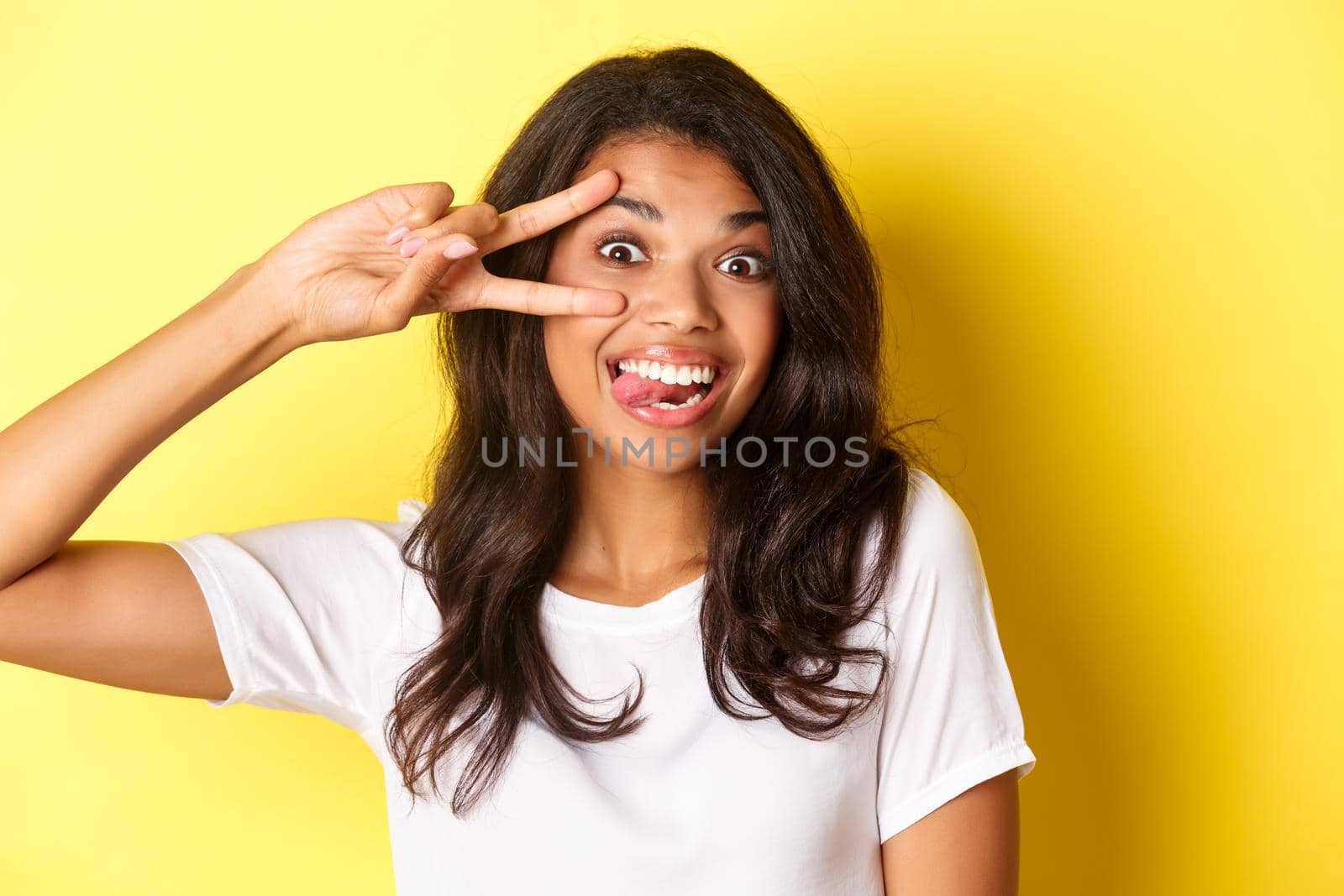 Image of silly and cute teenage african-american girl, showing peace sign and smiling, standing over yellow background.