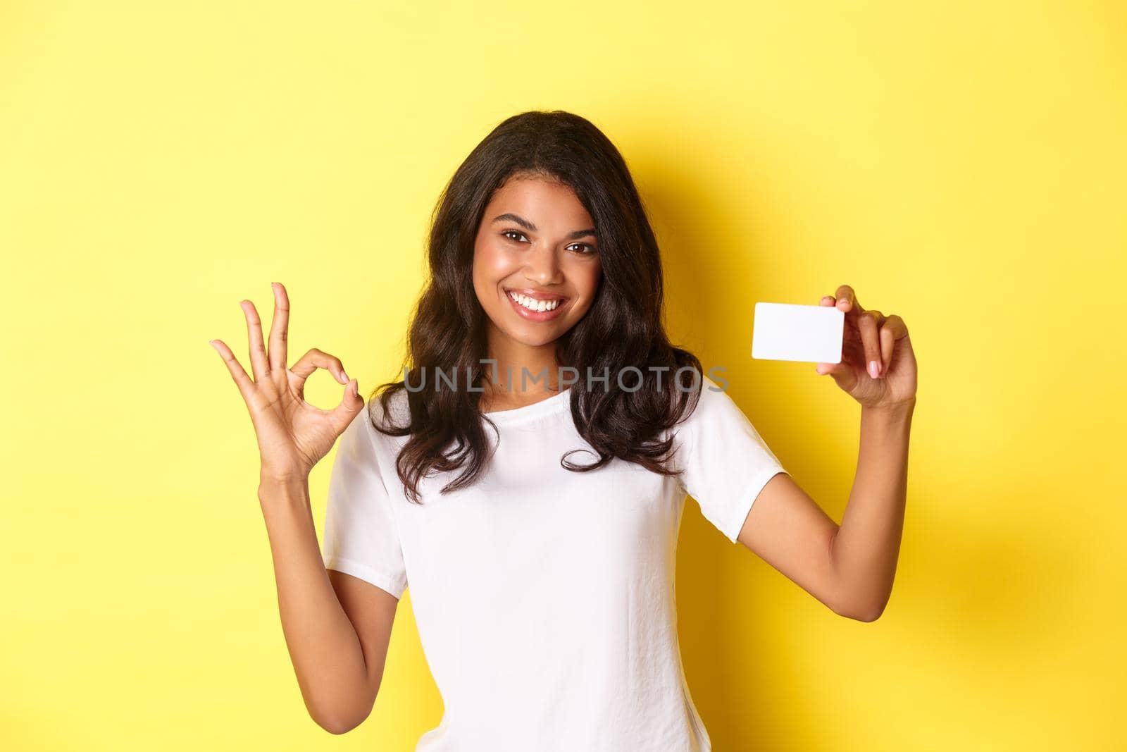 Portrait of young african-american girl showing credit card, making okay sign and smiling, recommending bank, standing over yellow background.