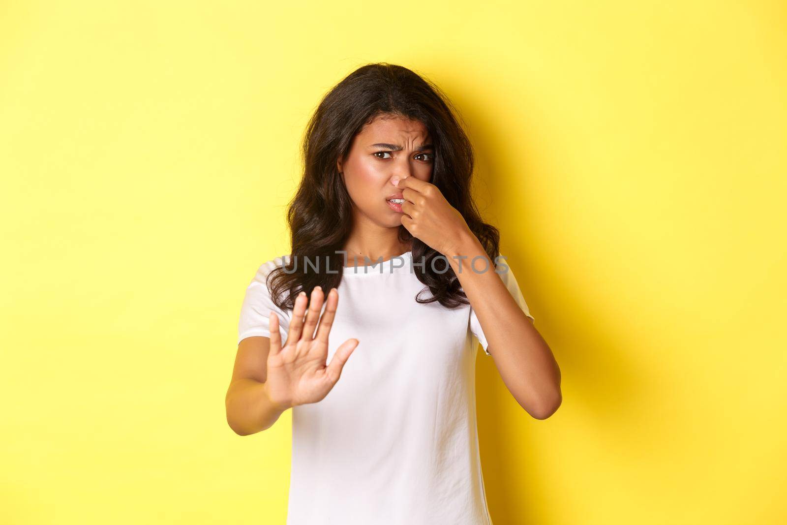 Young african-american woman in white t-shirt, shut her nose, looking disgusted and asking to stay away, dislike bad smell, standing over yellow background.