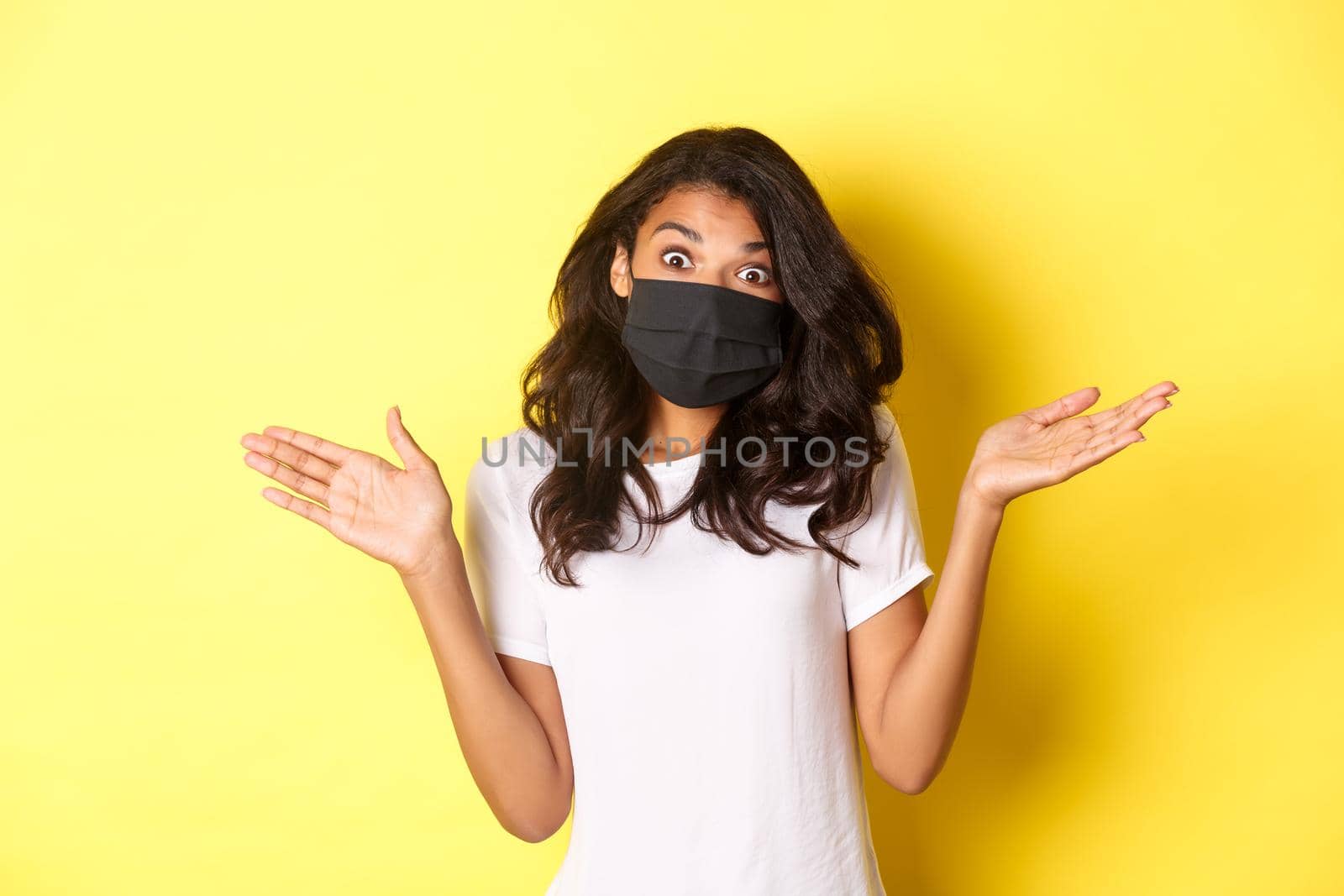 Concept of coronavirus, pandemic and lifestyle. Image of cute african-american girl in face mask, shrugging and looking clueless, dont know anything, standing over yellow background.