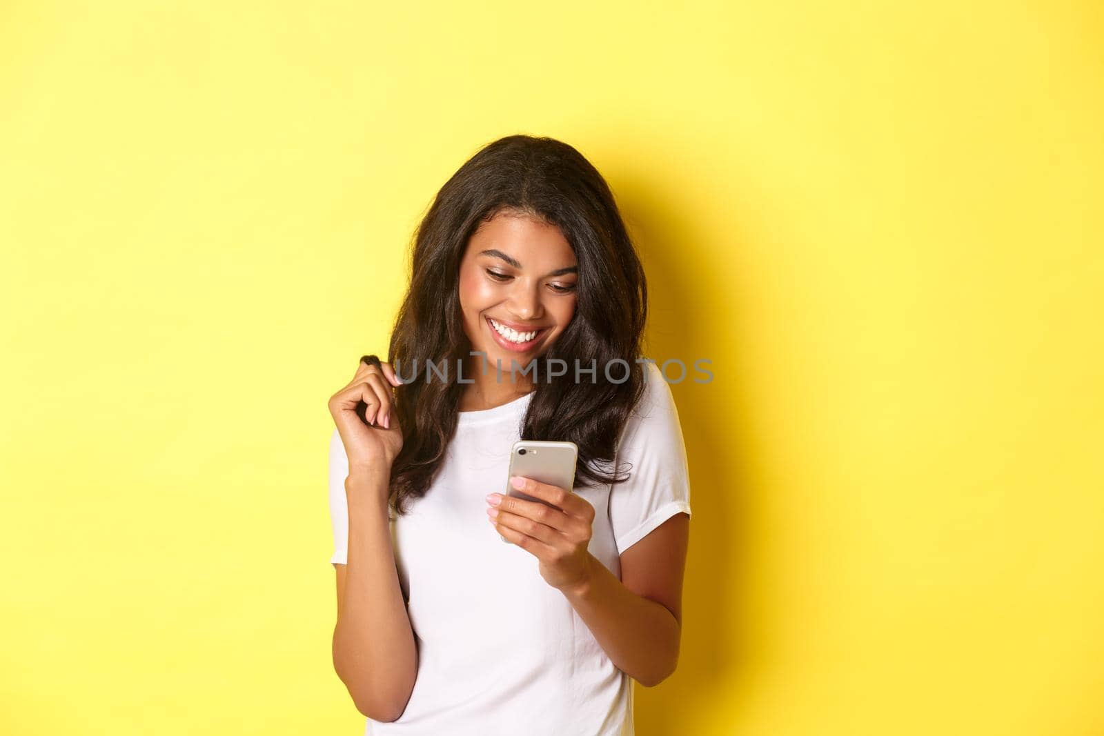 Image of attractive african american girl in white t-shirt, messaging on smartphone, looking at mobile phone and smiling, standing over yellow background by Benzoix