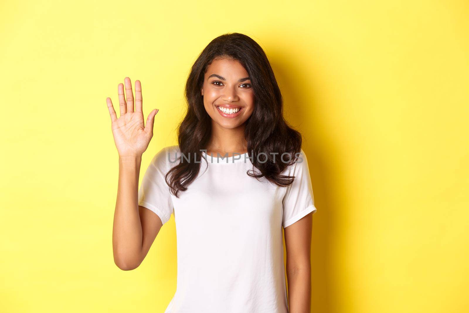 Image of beautiful and friendly african-american woman, waiving hand and smiling, saying hello, greeting you, standing over yellow background.