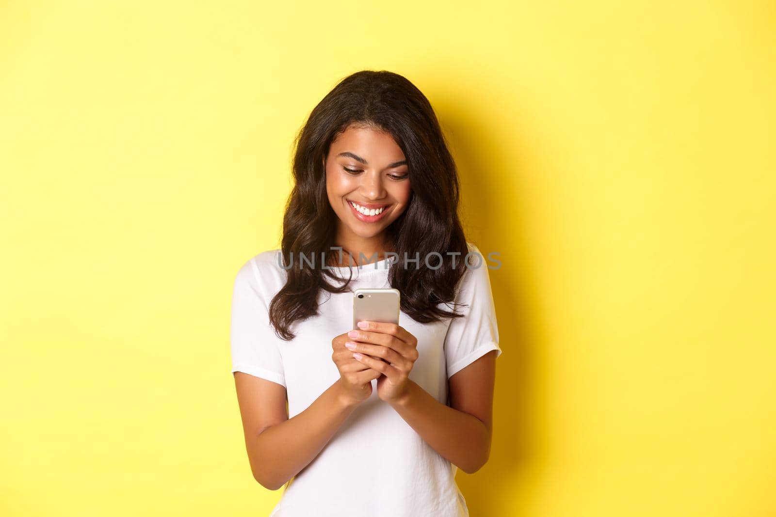 Portrait of good-looking african american girl in white t-shirt, messaging with smartphone, using mobile phone app, standing over yellow background by Benzoix