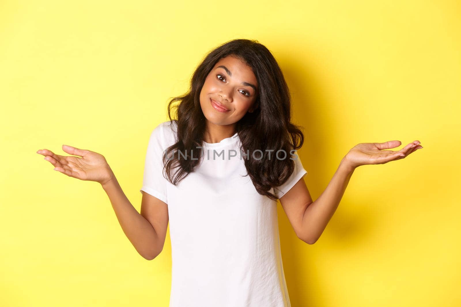 Portrait of clueless, beautiful african-american girl, shrugging and smiling, apologizing for being unaware, standing over yellow background.