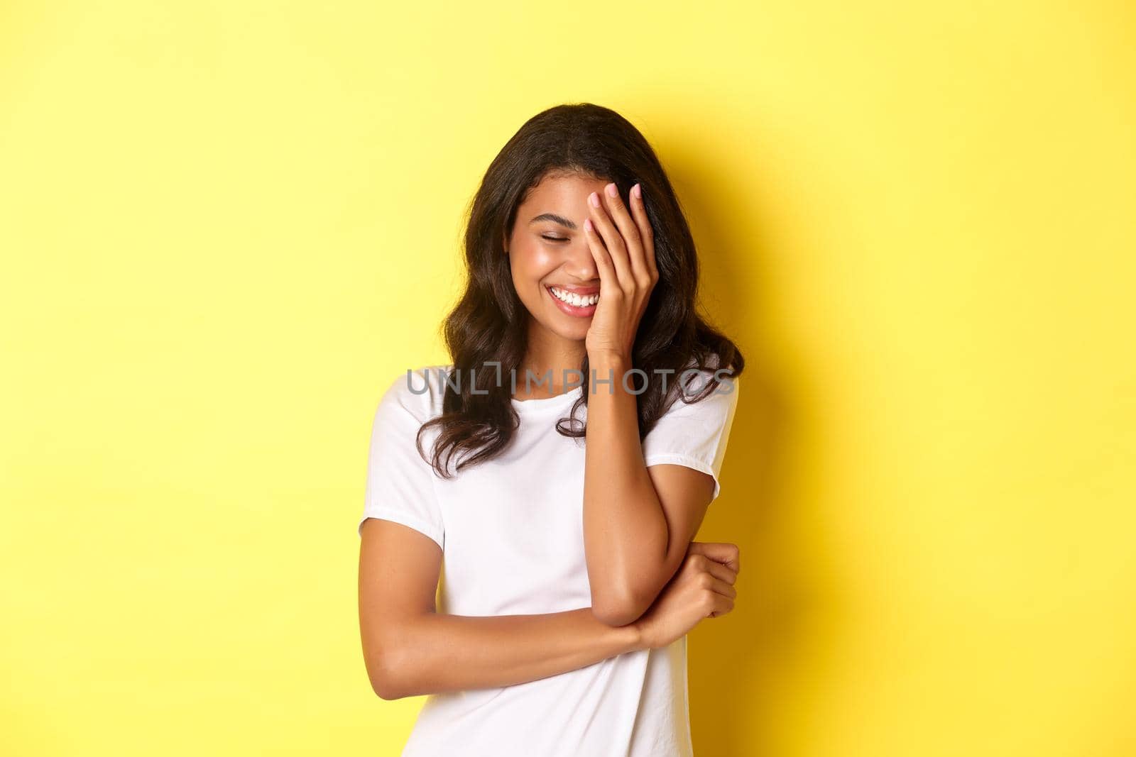 Portrait of beautiful and happy african-american girl, silly laughing and covering her face with hand, standing over yellow background by Benzoix
