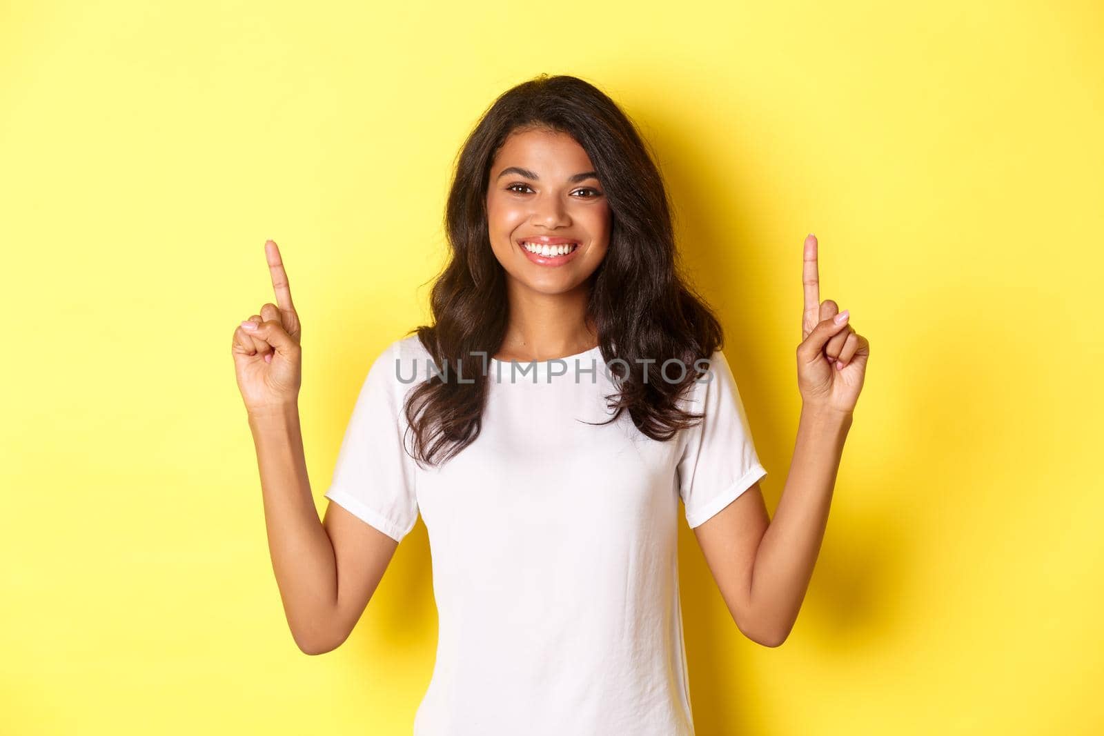 Portrait of attractive and confident african-american female model, wearing white t-shirt, pointing fingers up at promo logo, standing over yellow background by Benzoix