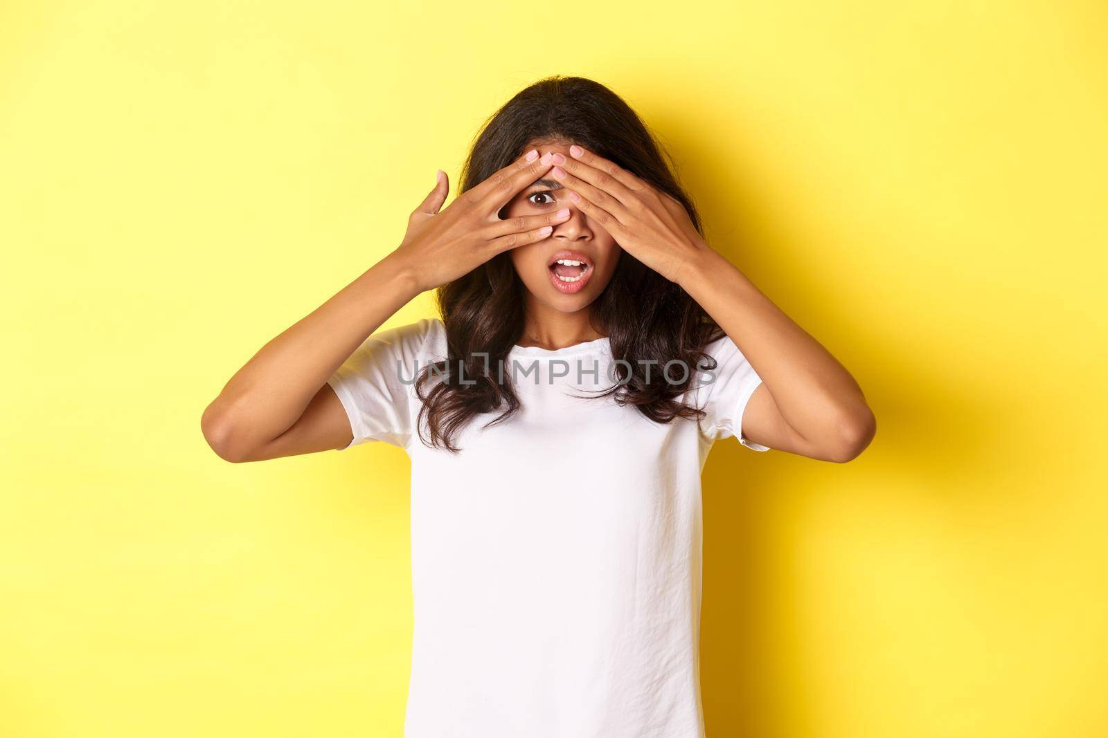 Portrait of embarrassed and shocked african-american girl, shut her eyes but peeking through fingers at something disturbing, standing over yellow background by Benzoix