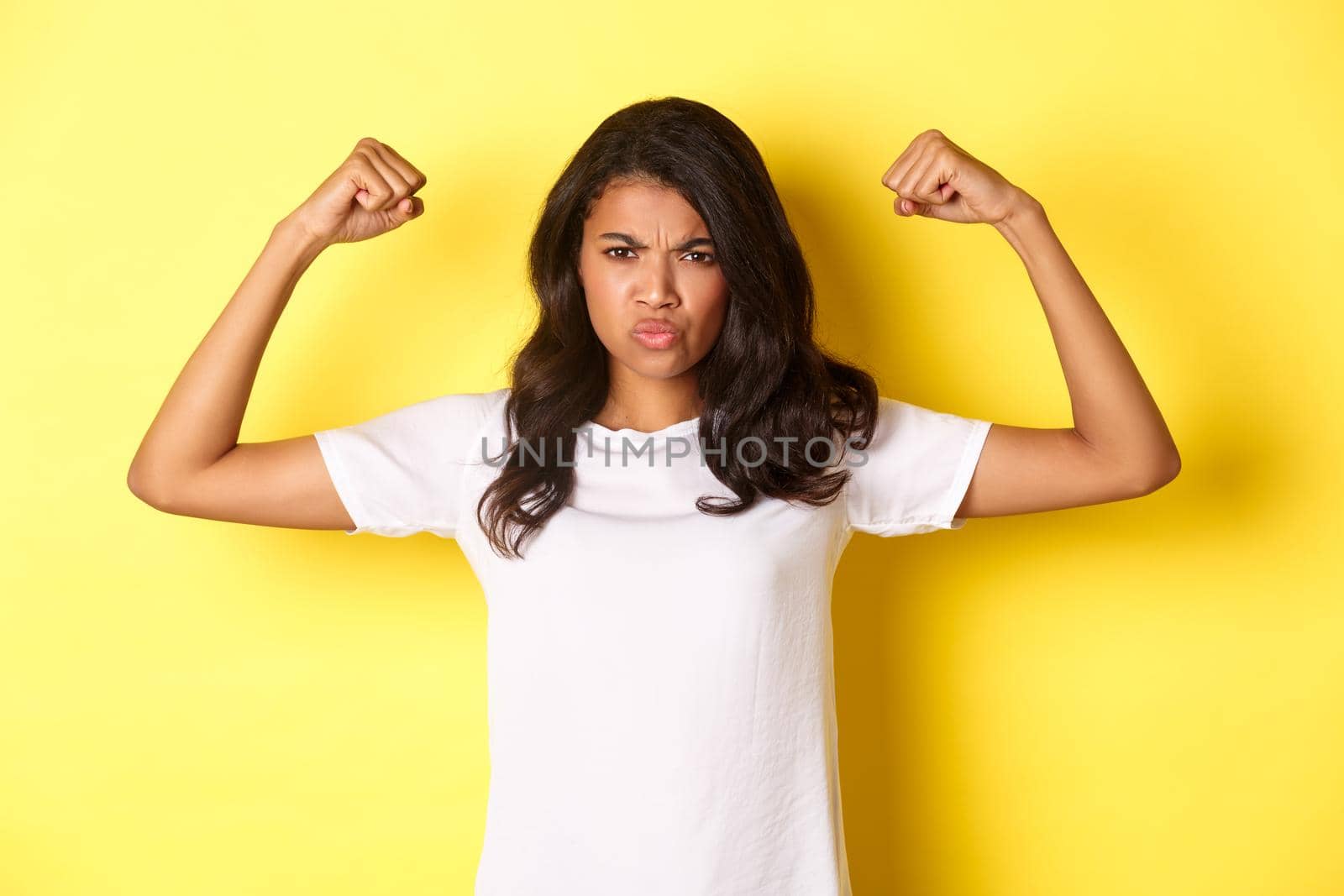 Image of strong and confident african-american girl, flexing biceps and looking self-assured, standing over yellow background.