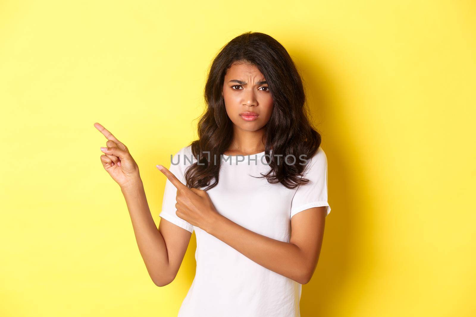 Portrait of displeased and worried african-american woman, expressing her doubts, pointing fingers at upper left corner and frowning hesitant, standing over yellow background by Benzoix