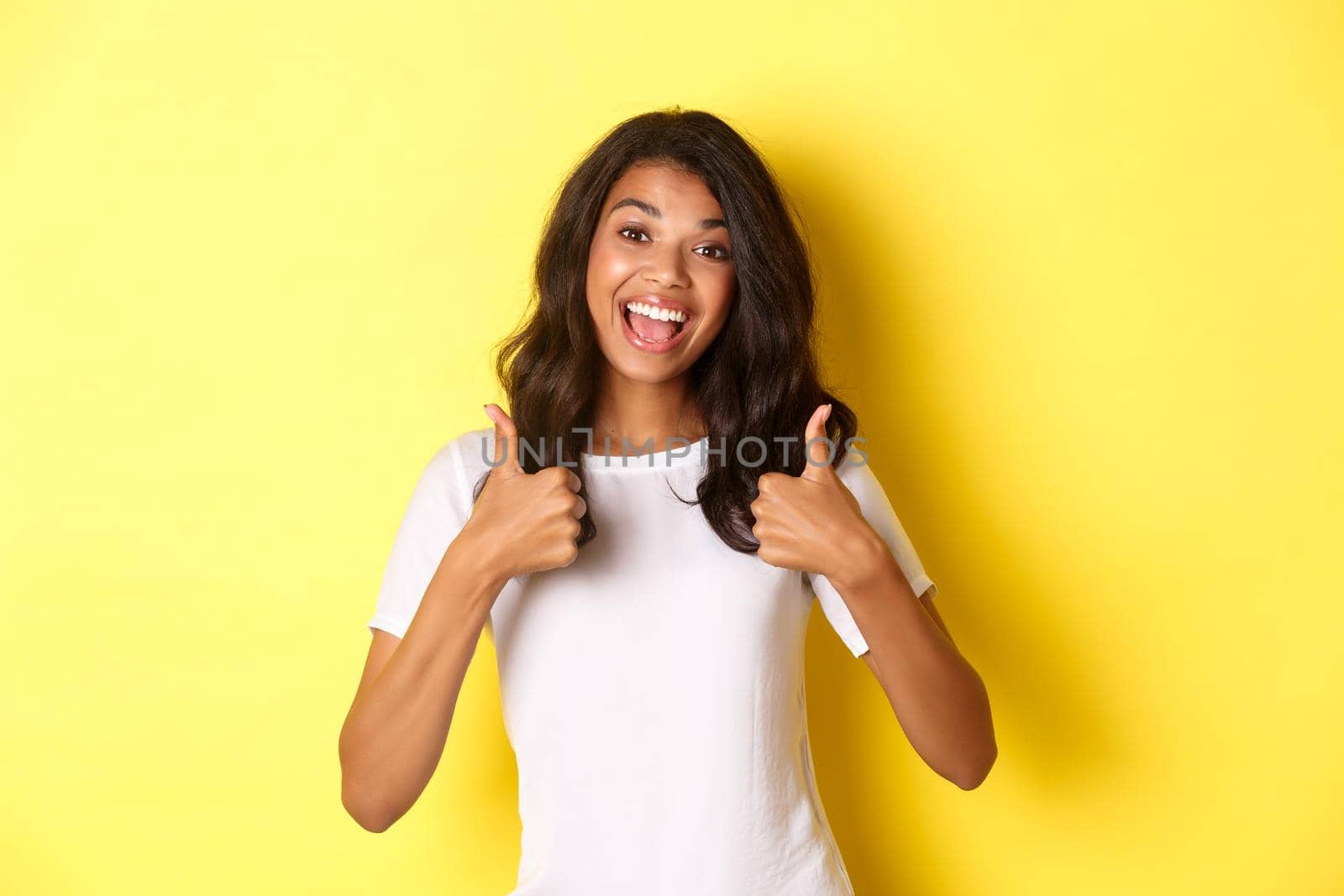 Portrait of cute and cheerful african-american girl, showing thumbs-up in support, like and agree with you, praising excellent work, standing over yellow background.