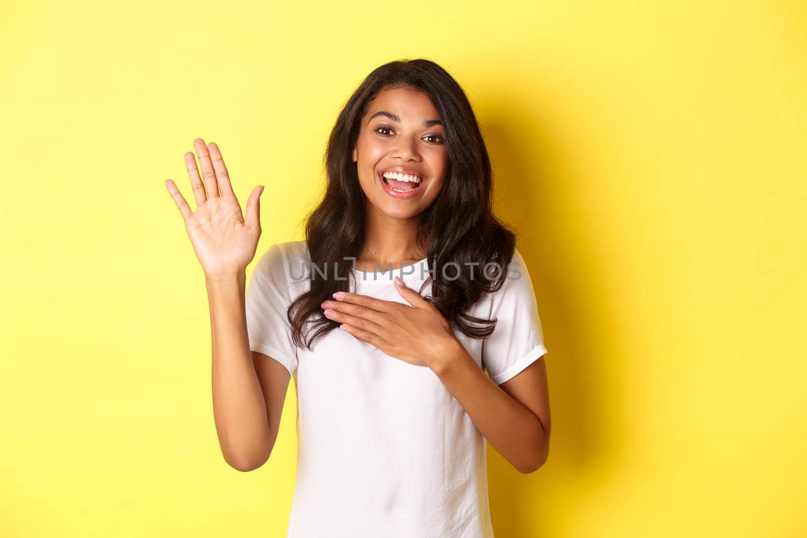 Portrait of cute and sincere african-american girl, making a promise, holding on hand raised and another on heart, swearing tell truth, standing over yellow background.