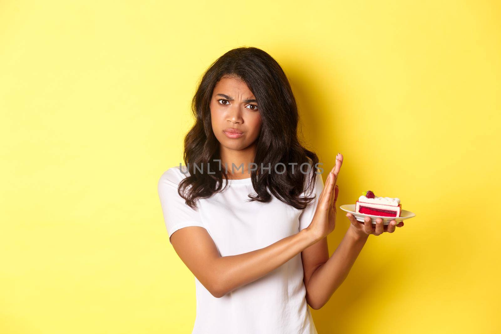 Image of displeased african-american girl decline to eat a cake, standing over yellow background.