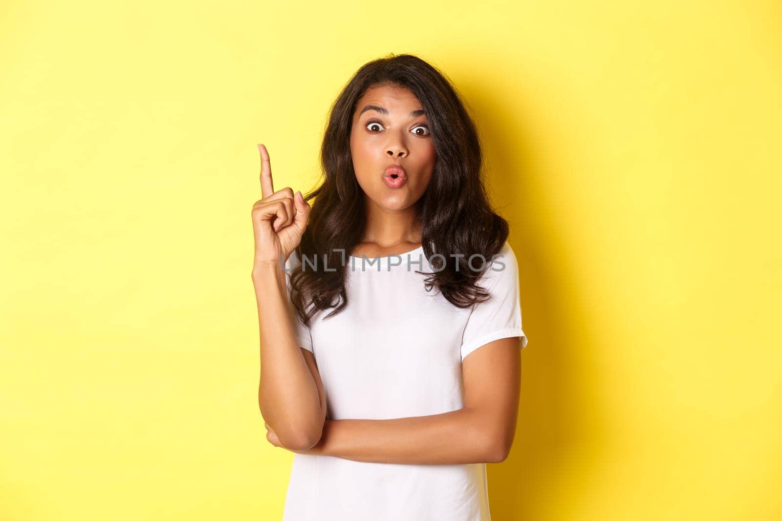 Portrait of smart african-american female student having a solution, suggesting idea, raising finger and looking excited, standing over yellow background by Benzoix