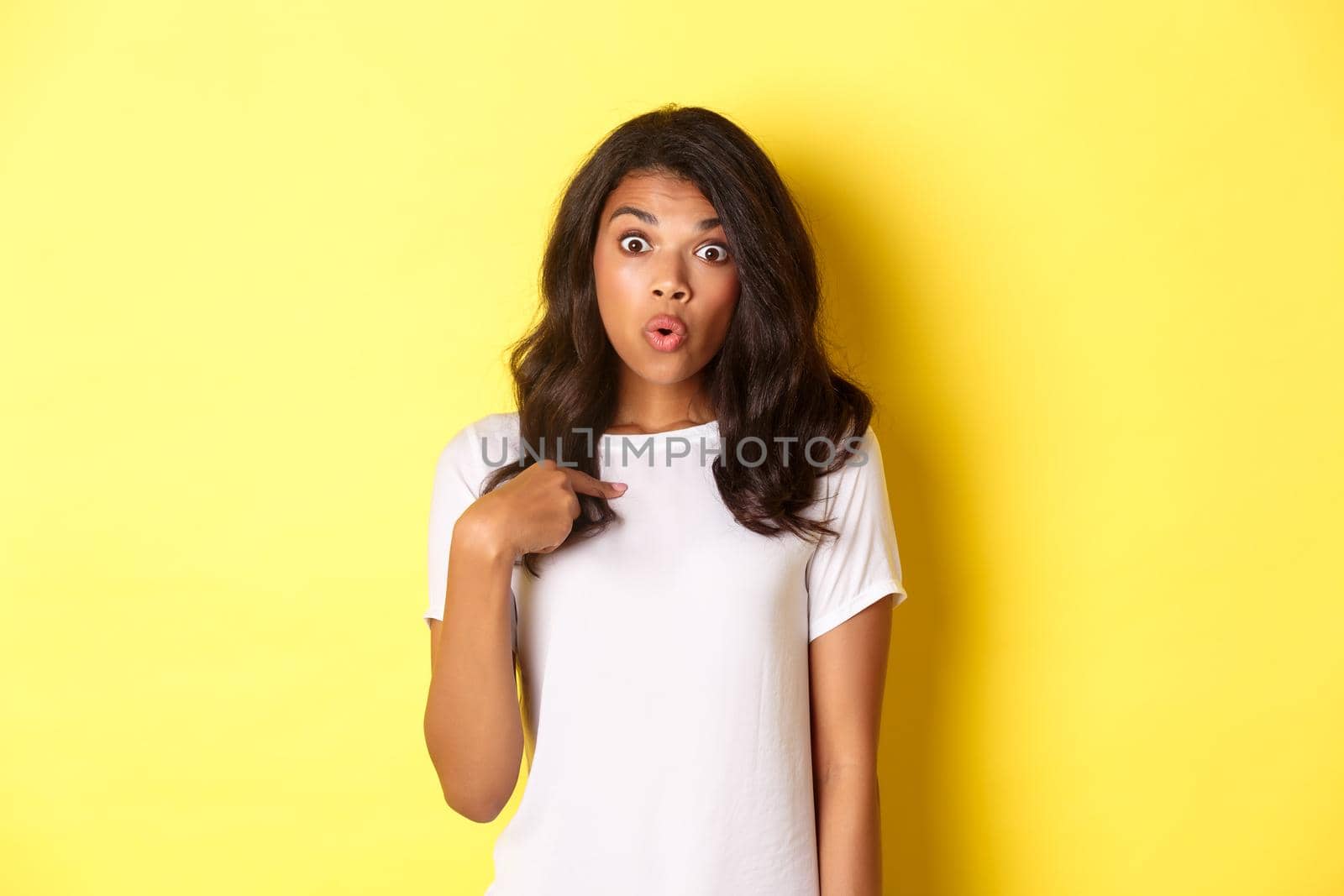 Portrait of surprised african-american girl, pointing finger at herself, standing over yellow background by Benzoix