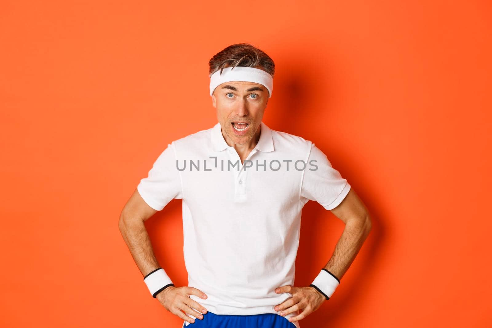 Concept of workout, sports and lifestyle. Portrait of startled middle-aged fitness guy in activewear, drop jaw and looking at camera amazed, standing over orange background.