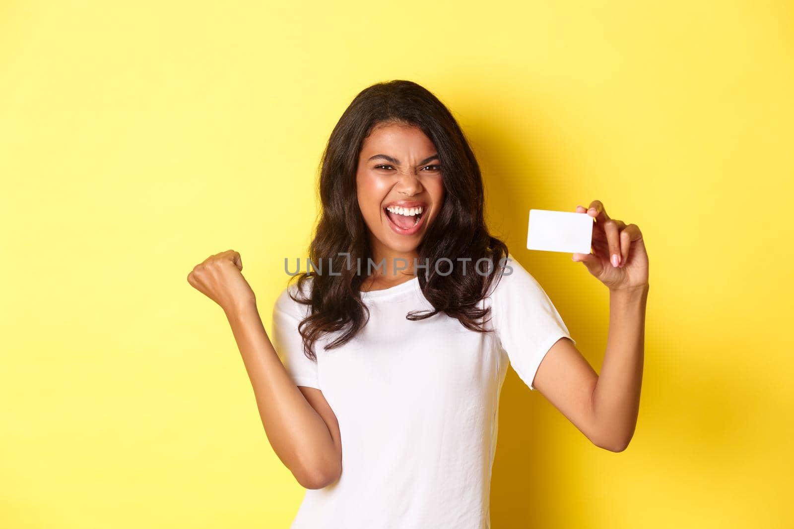 Portrait of cheerful african-american female model, showing credit card and shouting for joy, happy about shopping, standing over yellow background.