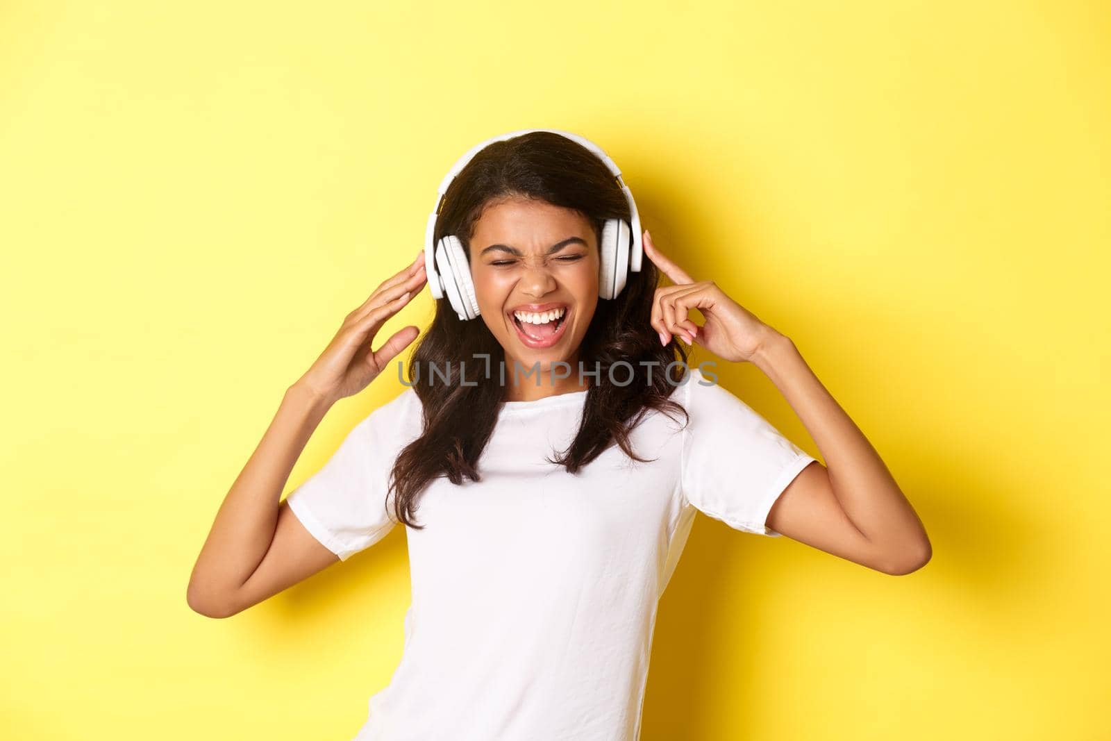 Portrait of modern african-american girl, enjoying listening to music in headphones, smiling amazed, standing over yellow background.