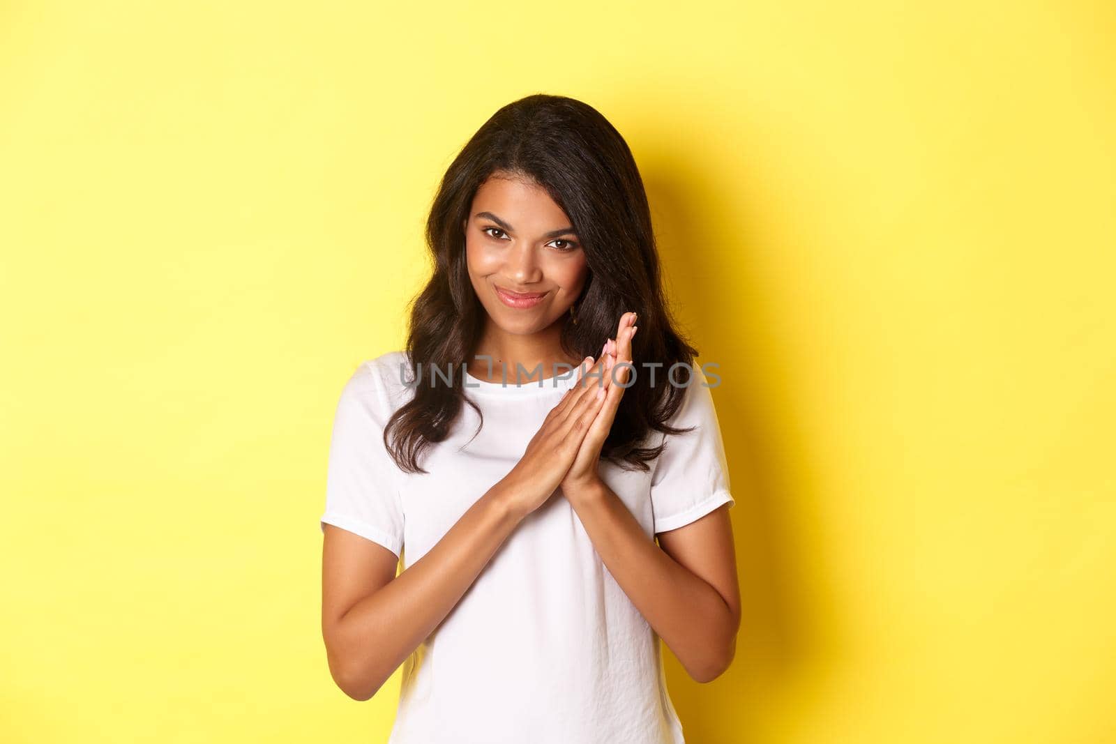 Image of cunning african-american girl, rubbing hands and smiling pleased, relish good deal, standing over yellow background.