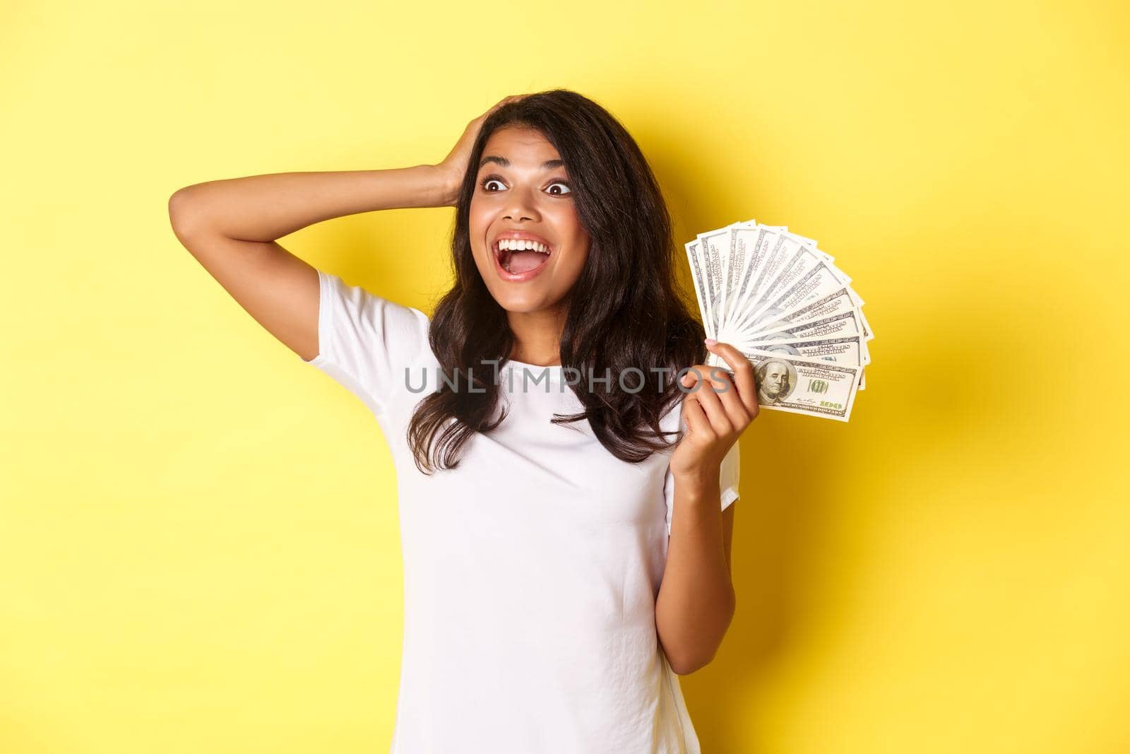 Image of lucky african-american girl, looking excited at upper left corner, holding money, going shopping, standing over yellow background by Benzoix