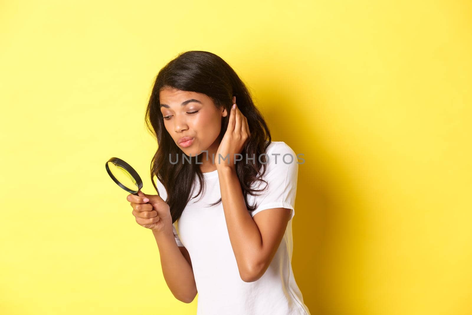 Image of cute african-american woman searching for something with magnifying glass, looking down, standing over yellow background.