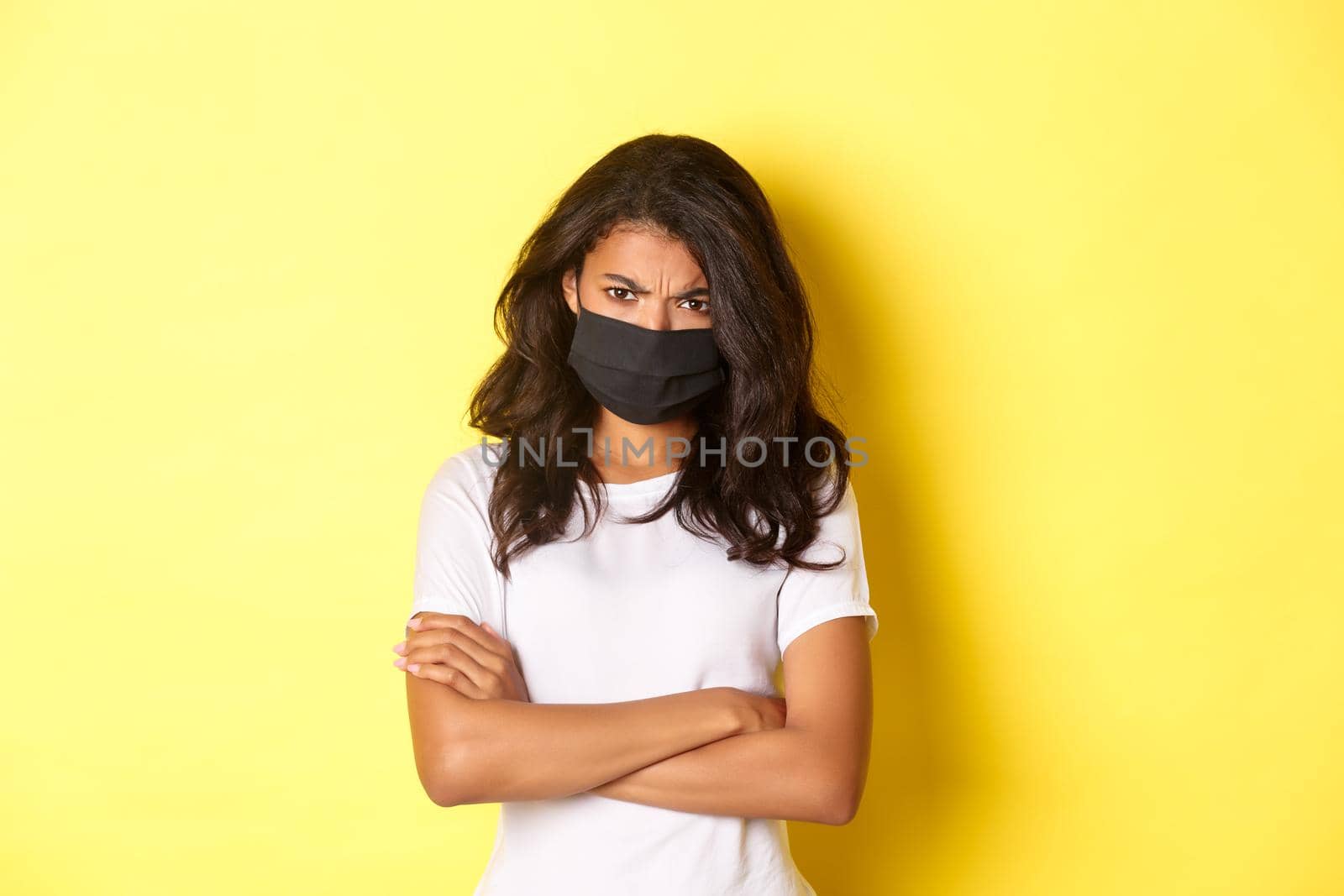 Concept of covid-19, social distancing and lifestyle. Angry and offended african-american girl, wearing face mask, being mad at someone, cross arms on chest and frowning, yellow background.