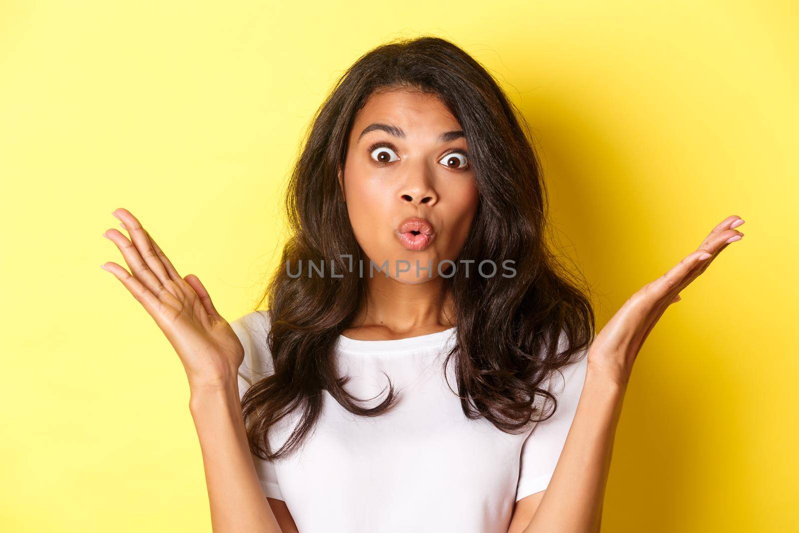 Close-up of surprised african-american girl, raising hands up and saying wow, hear amazing news, standing over yellow background.