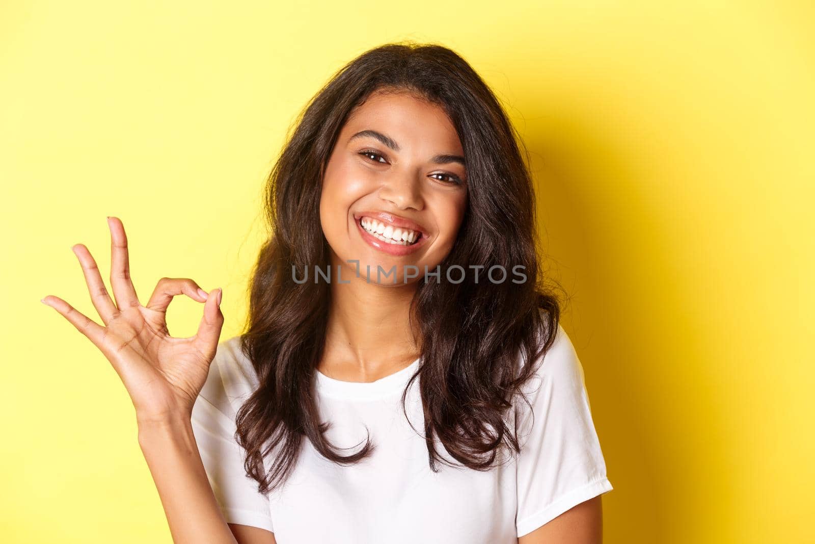 Close-up of satisfied african-american woman, showing okay sign and smiling, recommending something good, standing over yellow background by Benzoix