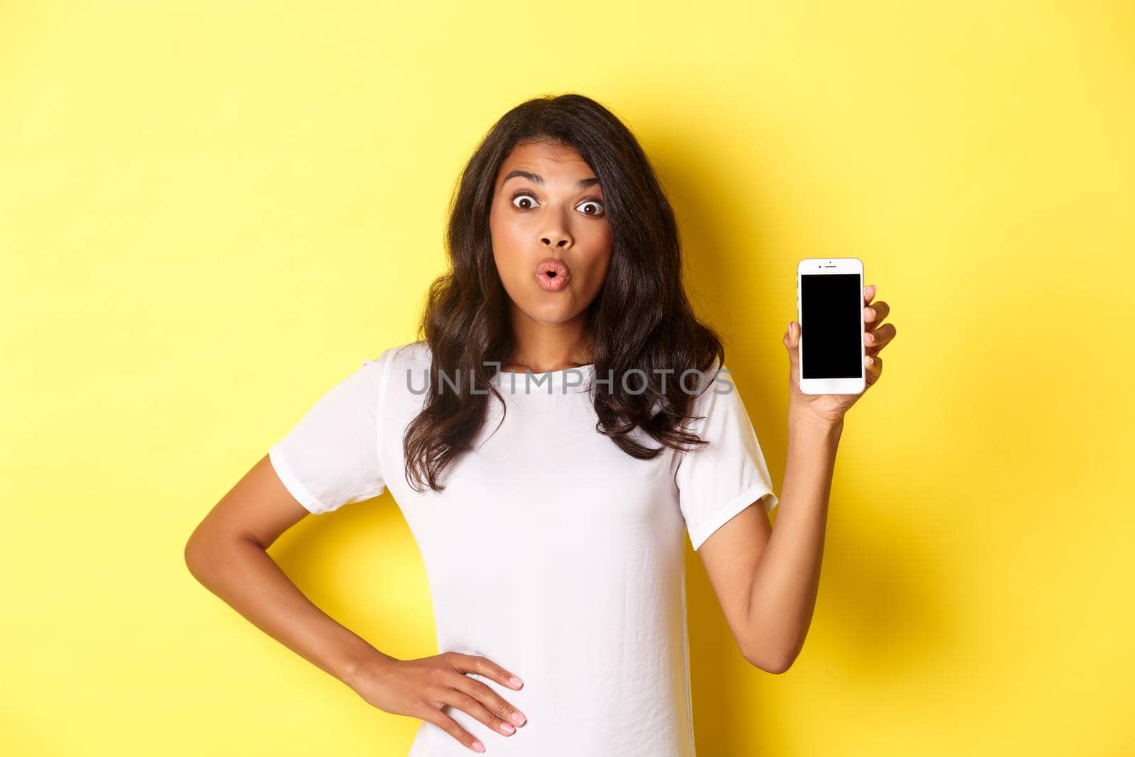 Image of amazed african-american girl, looking fascinated and showing smartphone screen, standing over yellow background by Benzoix