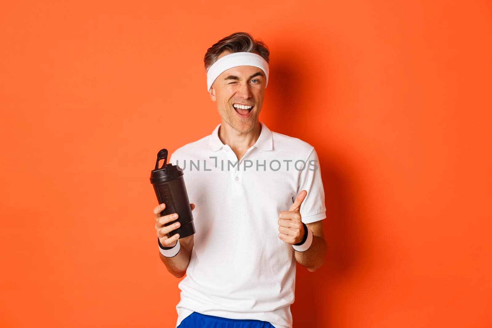 Portrait of confident, handsome middle-aged man in workout uniform, drinking water, winking and showing thumbs-up in approval, standing over orange background.