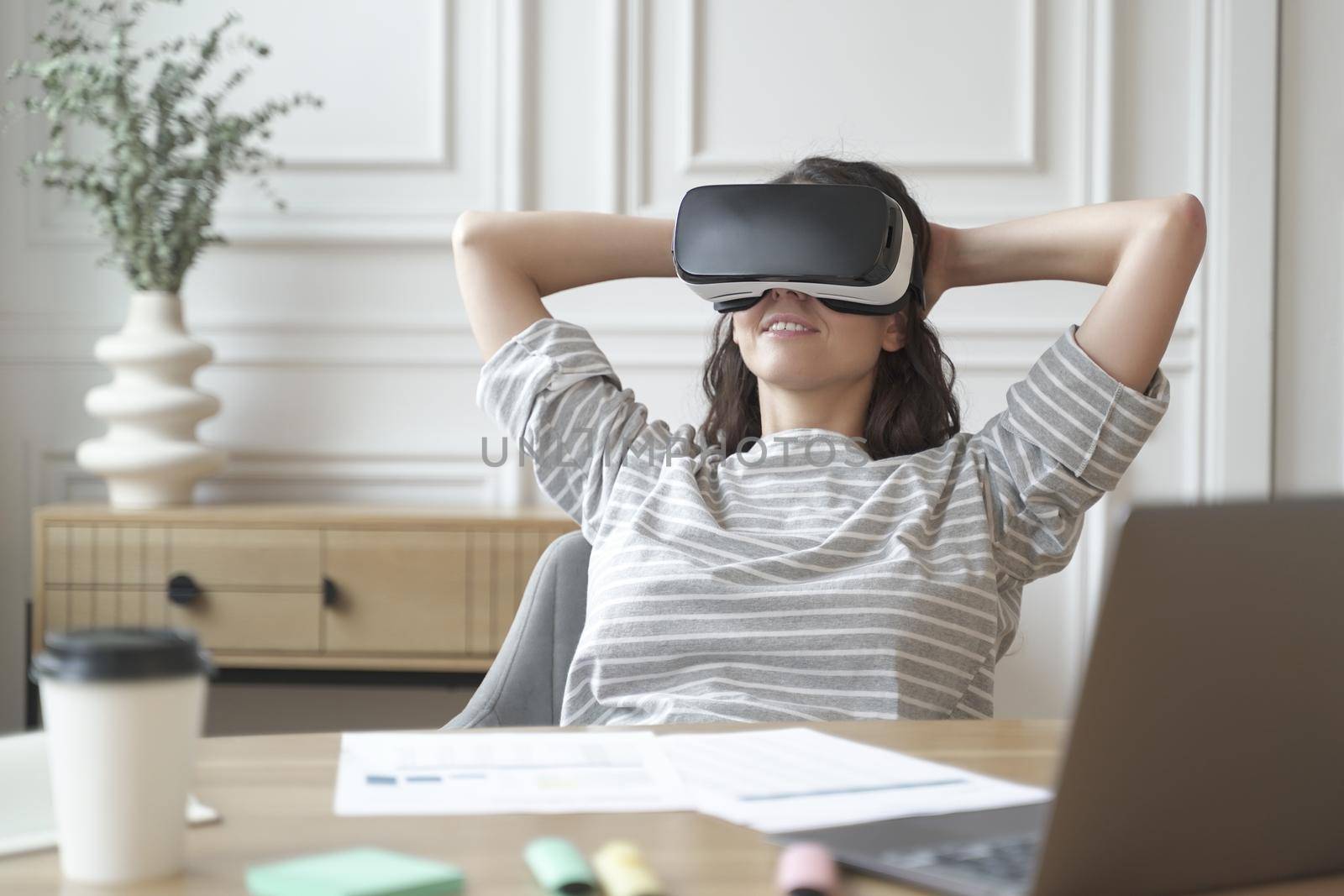 Relaxed young woman office worker wearing VR headset or helmet watching in 360 degrees video or movie during break at work, sitting in 3d glasses at workplace with hands behind head.