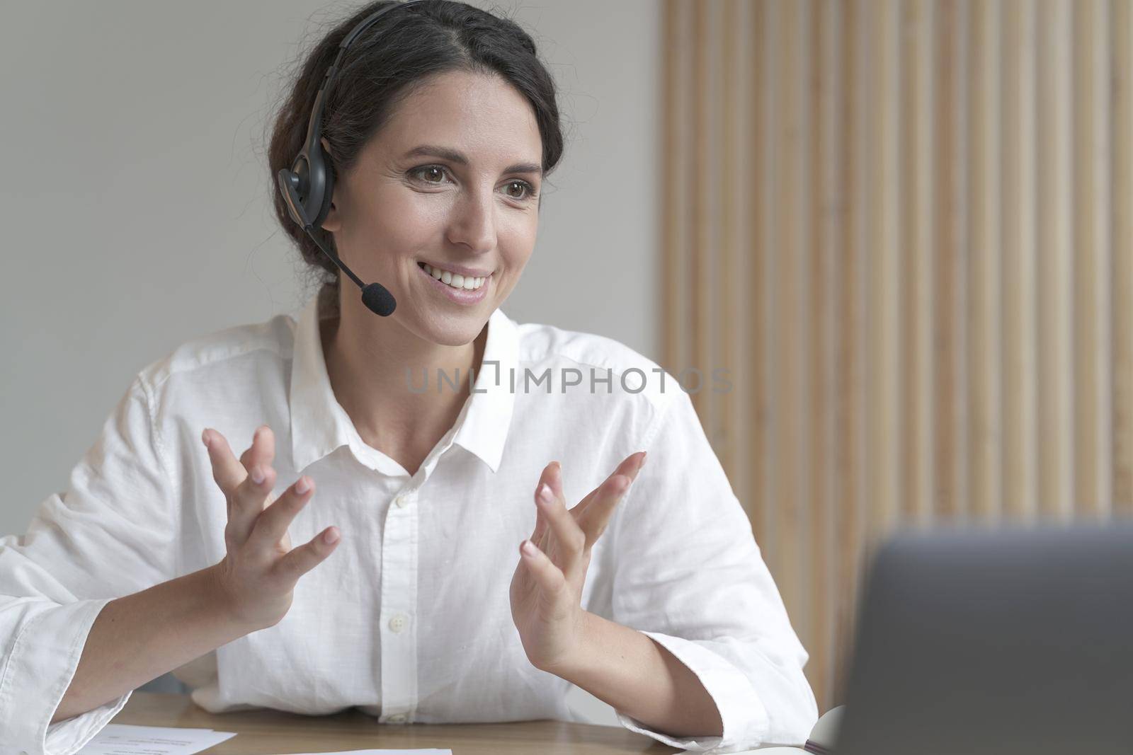 Head shot of pleasant young Italian female director wearing wireless headphones, looking at laptop screen, holding pleasant conversation with partners clients online, working remotely from home office