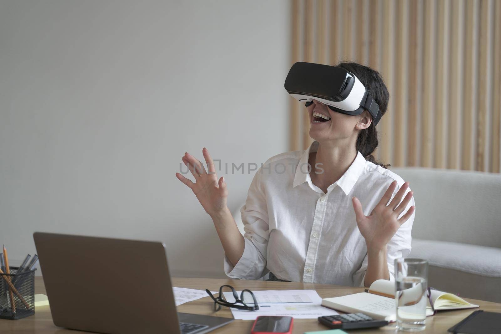 Enthusiastic business lady wears VR headset for laptop raising hands up as trying to touch objects in 3D reality when sitting at desk, working with apps online, modern technology for business concept
