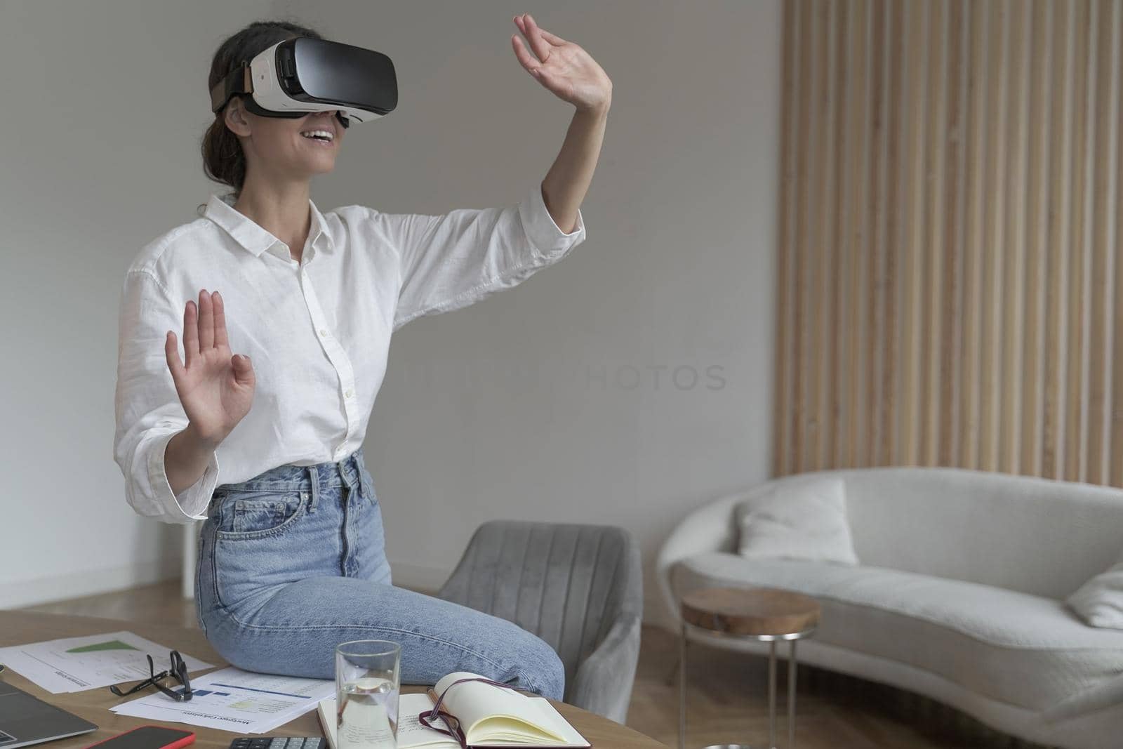 Joyful young business woman sitting on top of desk at home wearing VR glasses by vkstock