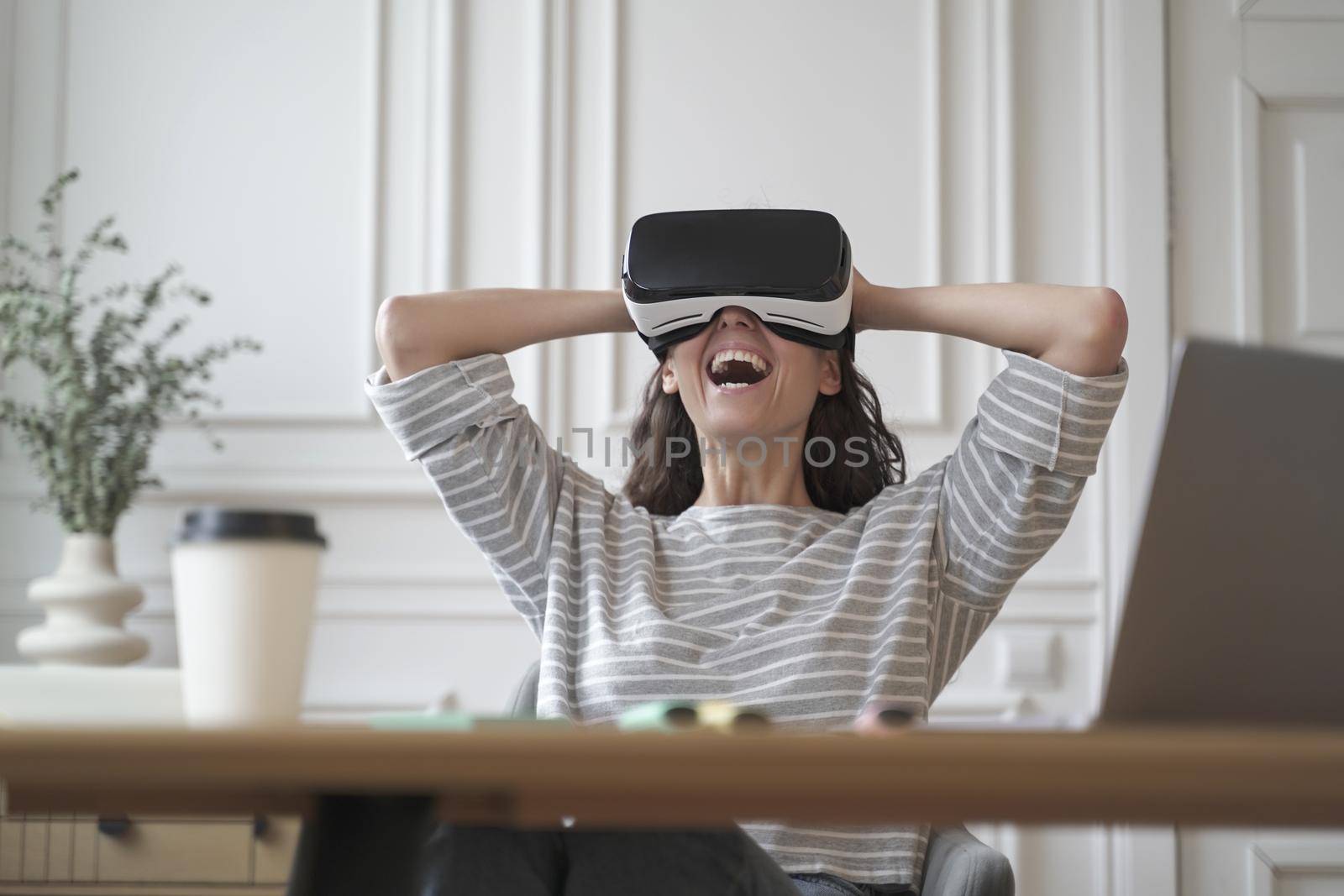 Amazed woman office employee in VR glasses laughing with joy leaning back in chair with hands behind head while remotely working from home office, cheerful lady spends free time in augmented reality
