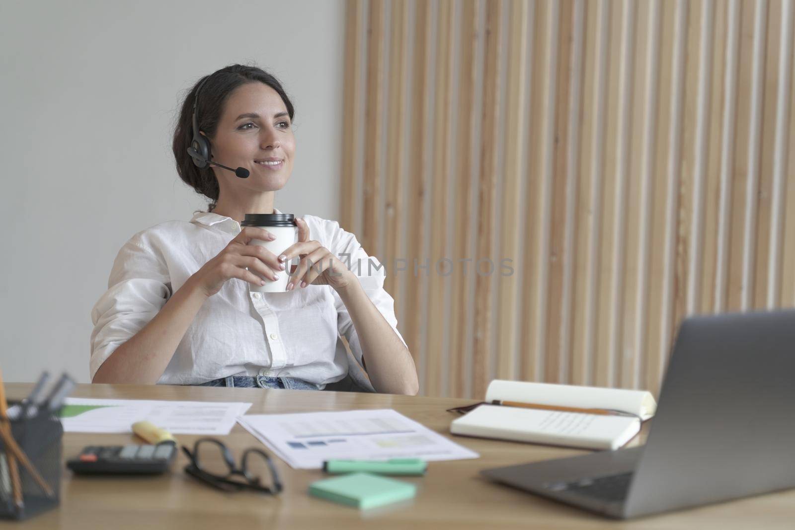 Pleasant italian business woman in headset sits at desk at home office with takeaway coffee by vkstock
