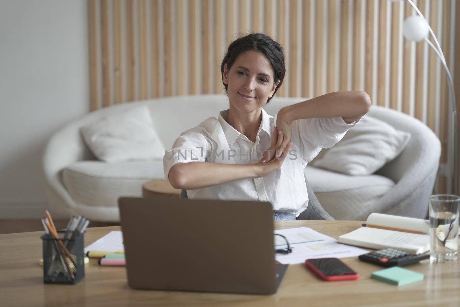 Positive business woman doing hands and fingers stretching exercises while listening to training lectures during webinar online on laptop. Freelancer lady broadly smiling as looking at computer screen