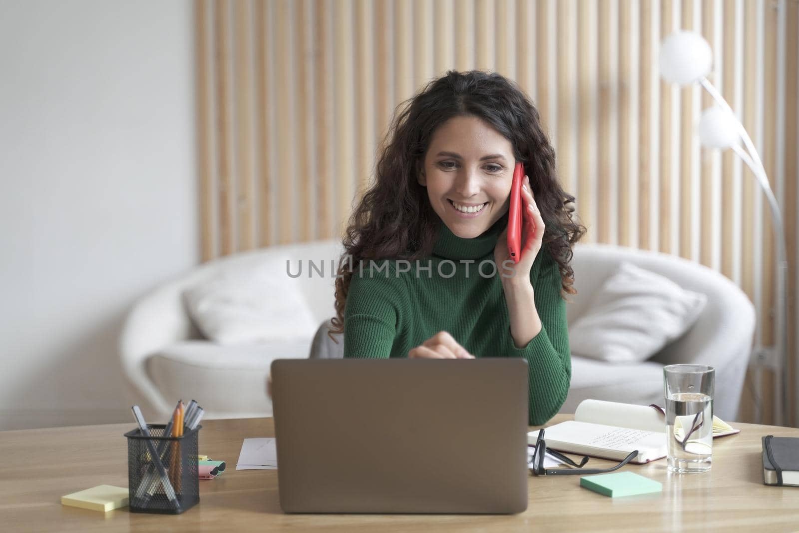 Happy successful Italian business woman having pleasant cellphone conversation while working on laptop at home office, smiling female employee discussing project with copartner by phone at work