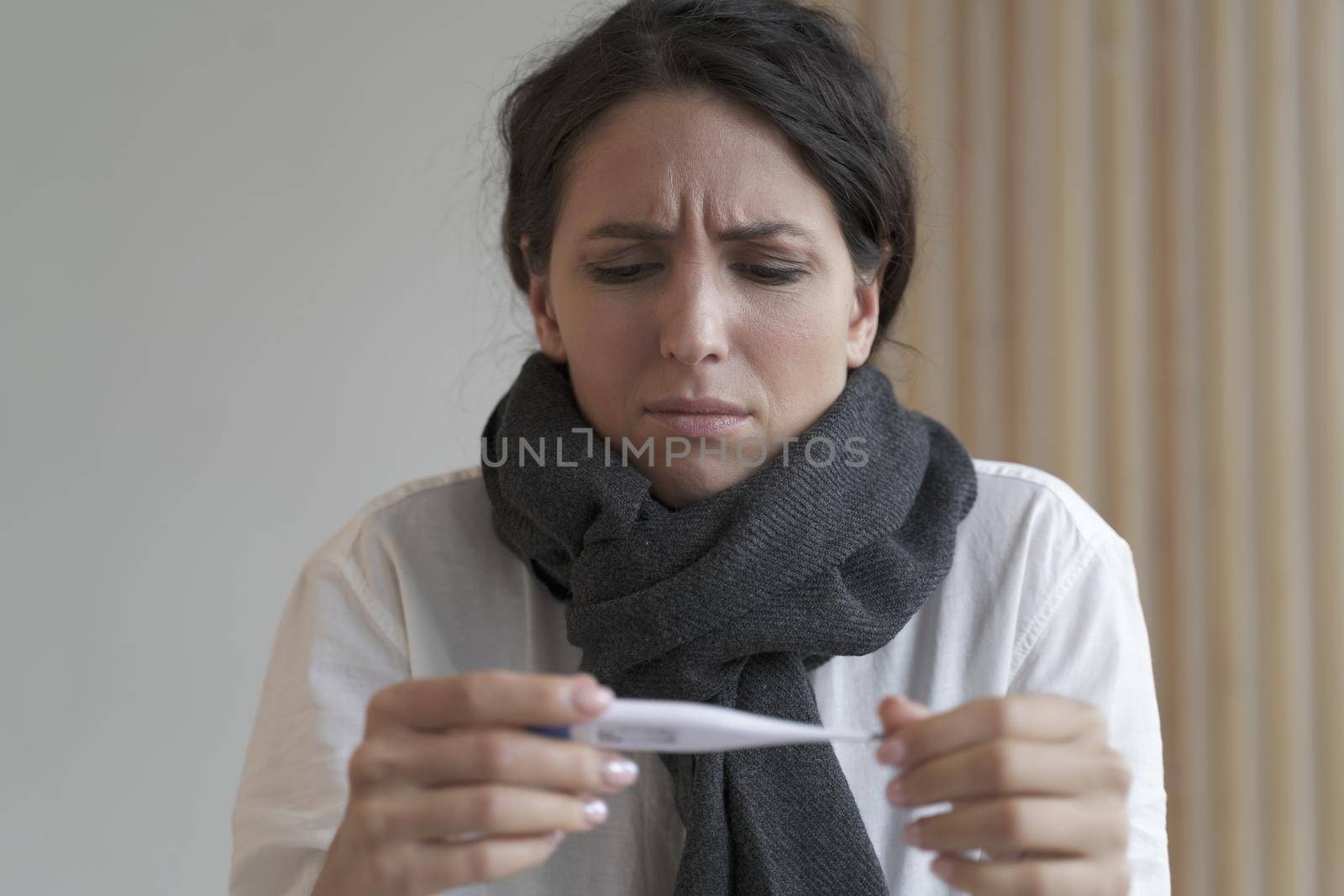 Sick-looking young business woman in knitted scarf looks frowning at electronic thermometer by vkstock
