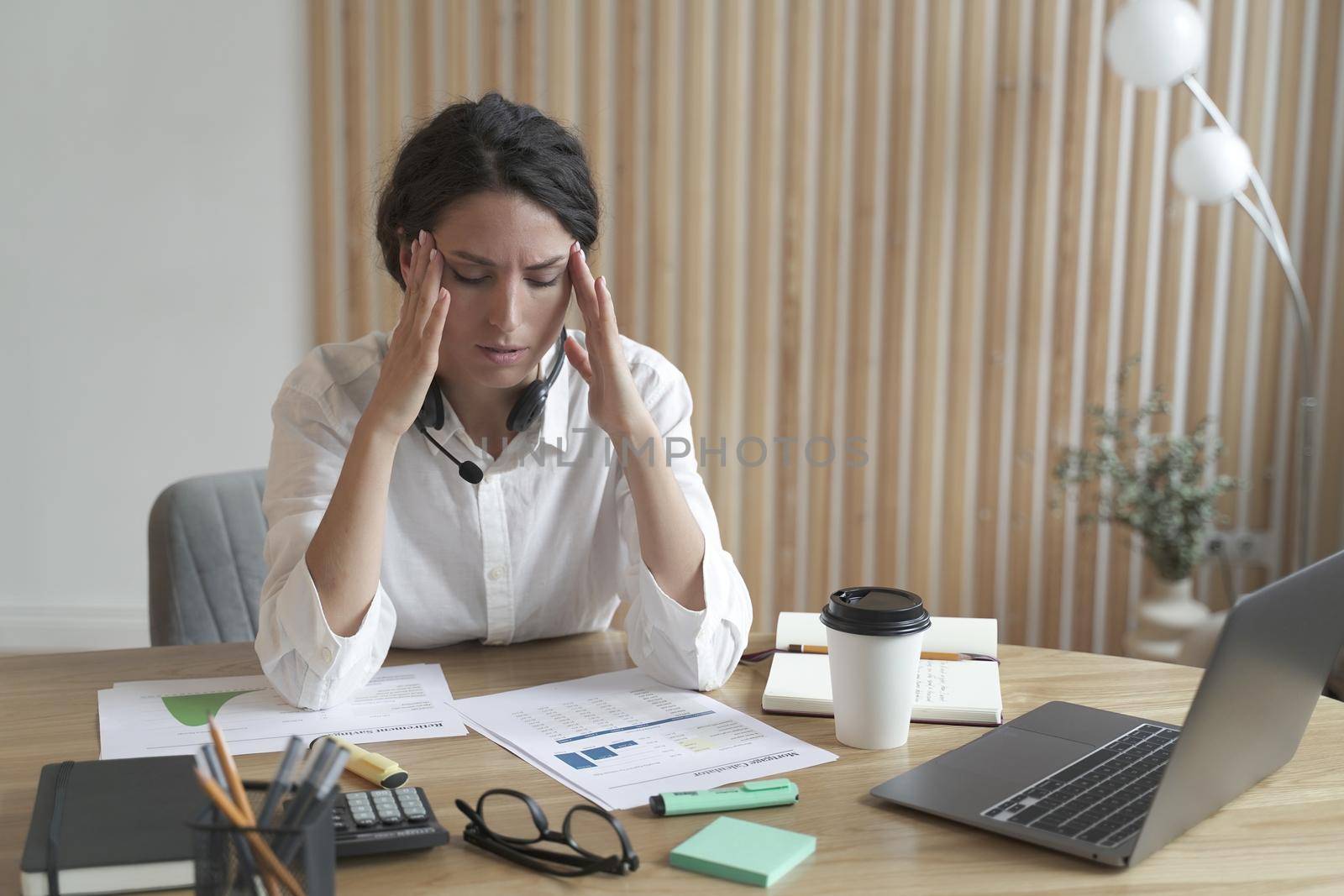 Young frustrated upset italian woman holding head in hands suffering from headache after hard working day, overworked sad business woman with closed eyes sitting at table with laptop and documents