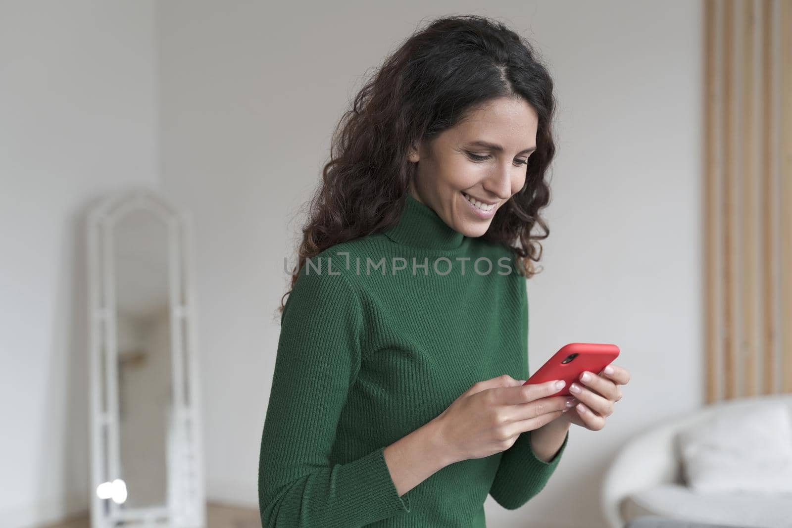 Young happy smiling Spanish woman using mobile phone and chatting with friends, texting sms or checking email while relaxing at home, pleased Italian female holding smartphone enjoying online shopping