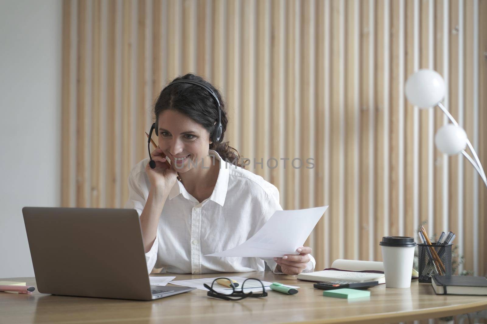 Happy young hispanic business woman in headset with microphone speaking by video call, looking at laptop computer while sitting at her workplace in modern office, call center agent consulting client