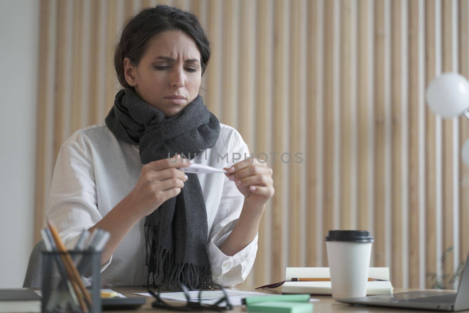 Unhealthy young hispanic business woman wearing scarf around neck holding thermometer, feeling unwell while working in office, female employee sitting at table and suffering of high fever