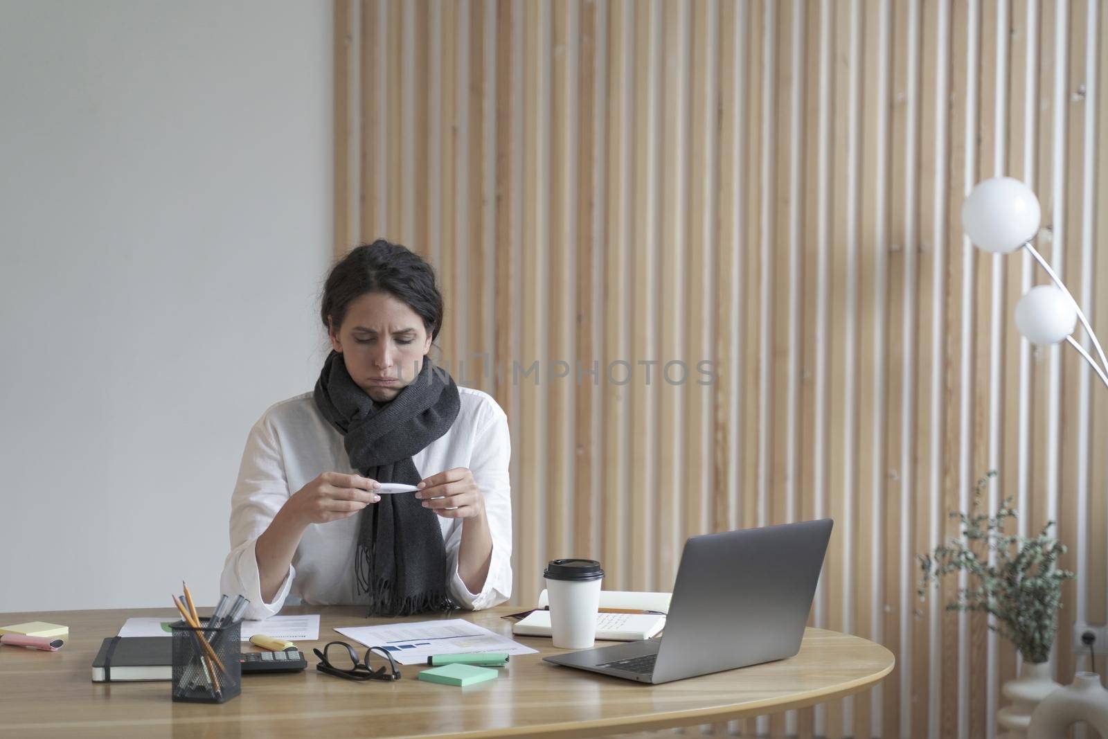 Unhappy sick young hispanic woman office worker in scarf around neck looking at electronic thermometer while sitting at her workplace, ill female suffering from covid19 or flu symptoms at work