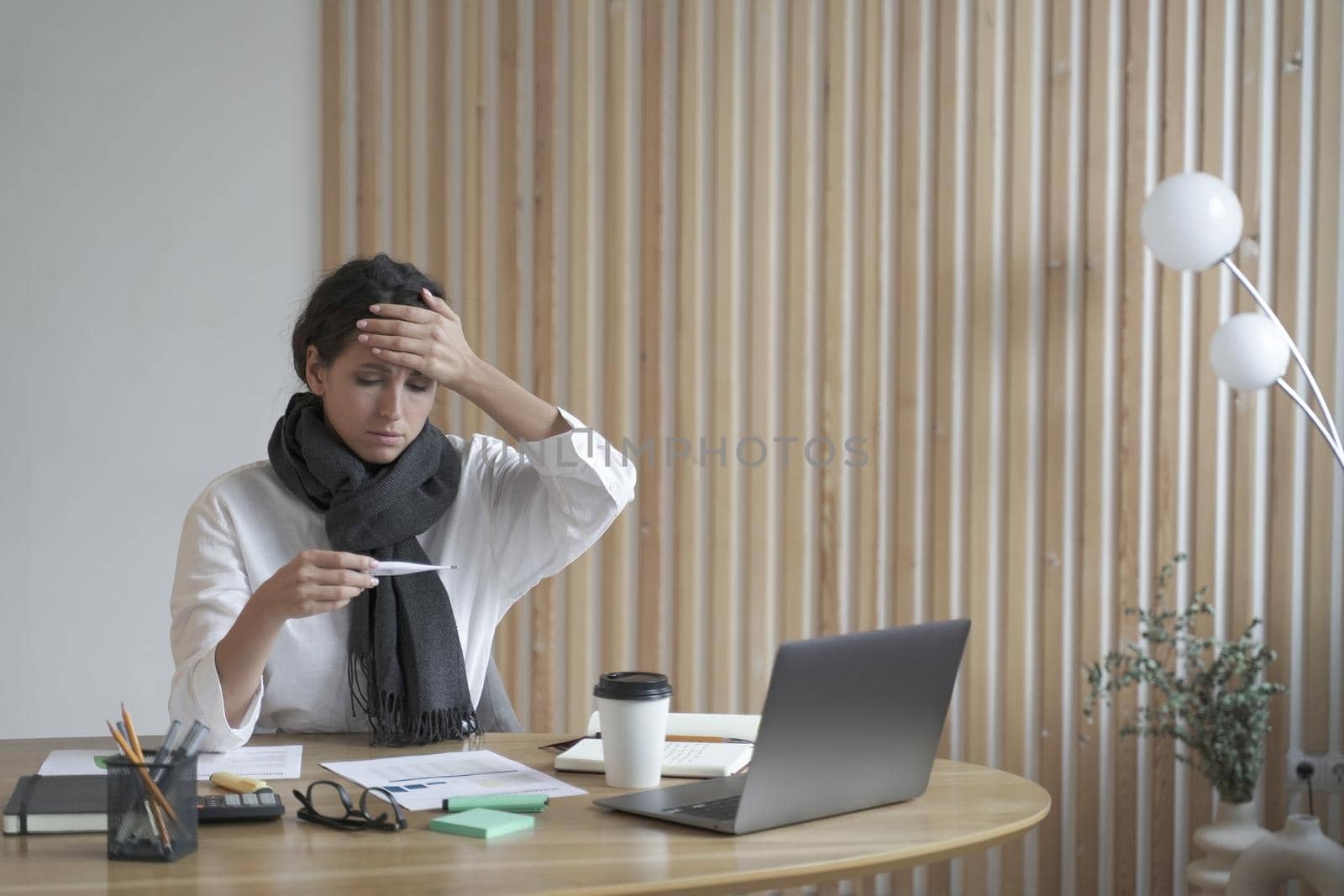 Tired lady freelancer sitting at desk at home office in front of laptop looking sickly at digital thermometer in her one hand while checking forehead with another. Bushed young woman overworked on pc